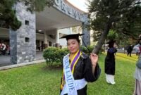 Seorang wisudawati berpose bahagia mengenakan toga lengkap dengan selempang predikat kelulusan 