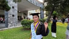 Seorang wisudawati berpose bahagia mengenakan toga lengkap dengan selempang predikat kelulusan 