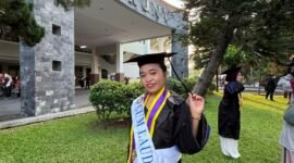 Seorang wisudawati berpose bahagia mengenakan toga lengkap dengan selempang predikat kelulusan 