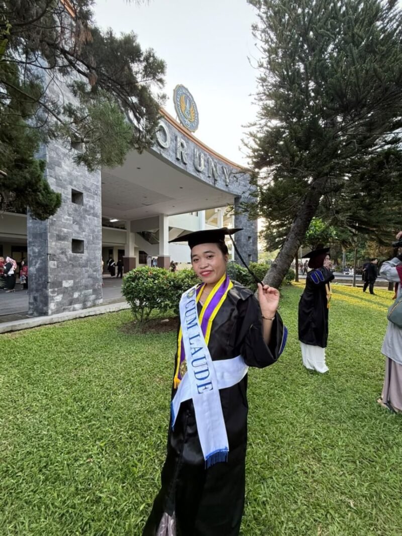 Seorang wisudawati berpose bahagia mengenakan toga lengkap dengan selempang predikat kelulusan 